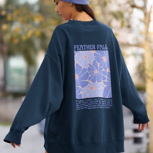 Puede incluir: Una sudadera azul marino con un diseño floral y el texto "FEATHER FALL" en la espalda. El texto debajo del diseño dice "La velocidad de descenso de una criatura que cae se reduce a 50 pies por ronda hasta que termina el hechizo. Si la criatura aterriza antes de que termine el hechizo, no recibe daño por caída y puede aterrizar de pie, y el hechizo termina para esa criatura."