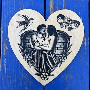 Könnte beinhalten: Herzförmiges Holzkunstwerk mit einem schwarz-weißen Tattoo-Design. Das zentrale Bild zeigt ein küssendes Paar innerhalb einer Backsteinmauer und eines floralen Randes. Eine Schwalbe und eine Motte sind ebenfalls enthalten.