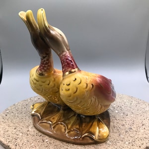 May include: A pair of ceramic ducks with yellow, brown, and red paint. The ducks are facing each other with their necks bent. They are standing on a brown base.