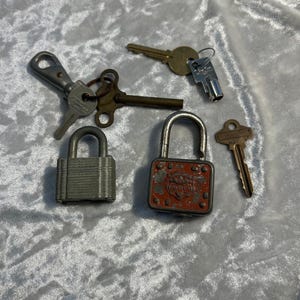 Puede incluir: Un surtido de llaves y candados antiguos. La colección incluye una variedad de estilos de llaves, incluyendo llaves esqueléticas y diseños modernos. Se ven dos candados, uno gris y otro rojo con la palabra "Master" en él. Los artículos están dispuestos sobre una superficie texturizada de color claro.