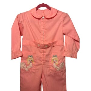 May include: A light pink jumpsuit with a Peter Pan collar and long sleeves. The jumpsuit features a front zipper, a belt, and two front pockets. Each pocket has an embroidered design of a blonde girl with a squirrel body. The jumpsuit is hanging on a pink hanger.