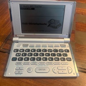 Peut inclure: Un dictionnaire électronique Casio argenté avec clavier et écran. L'écran affiche "Multi-Wörterbuchsuche" et d'autres textes. L'appareil possède diverses touches de fonction et une disposition de clavier QWERTY. Le numéro de modèle est EW-G2300.