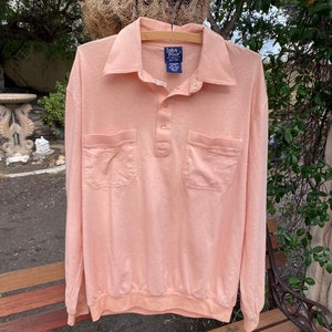 May include: A peach-colored long-sleeved polo shirt with a button-down collar and two patch pockets. The shirt has the text "John Blair" on the tag.