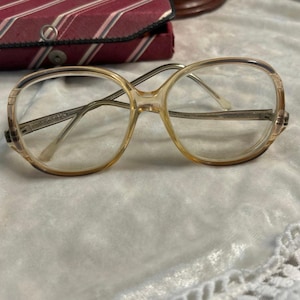 May include: Vintage eyeglasses with a light brown frame and clear lenses. The glasses have a rounded shape and a metal bridge. A striped case is in the background.