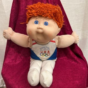 May include: A plush doll with bright orange yarn hair and blue eyes, wearing a white and blue tank top featuring the text "OlympiKids 1996 Official Team Mascot USA" and the five-ring Olympic logo, paired with white pants and soft fabric limbs.