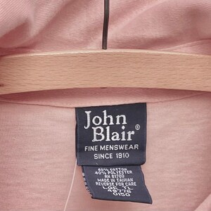 May include: A black tag with white text that reads "John Blair Fine Menswear Since 1910". The tag is attached to a pink shirt. The tag also includes the following information: 60% Cotton, 40% Polyester, RN 81700, Made in Taiwan, Reverse for Care, LOE TL 46776 0150.
