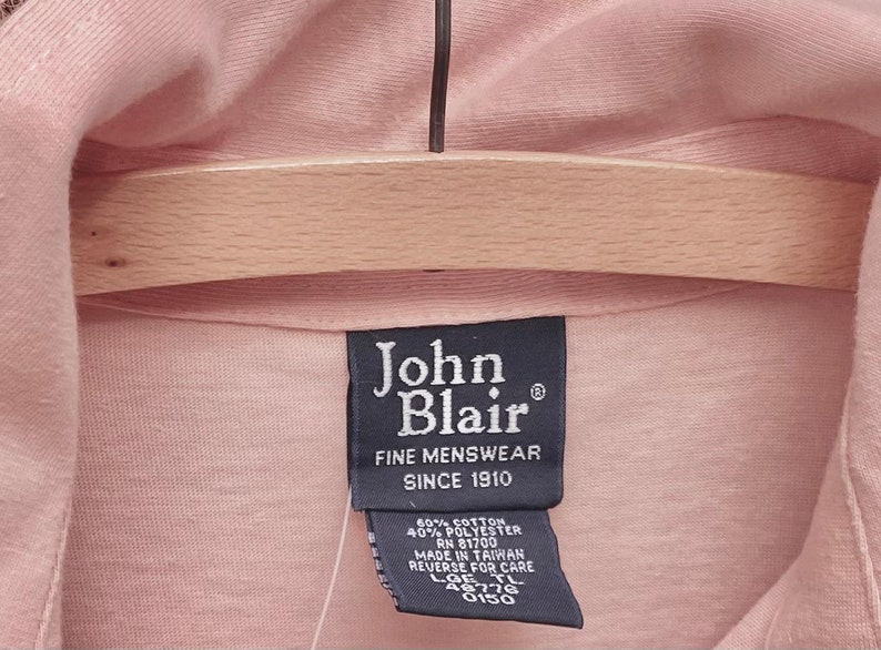 May include: A black tag with white text that reads "John Blair Fine Menswear Since 1910". The tag is attached to a pink shirt. The tag also includes the following information: 60% Cotton, 40% Polyester, RN 81700, Made in Taiwan, Reverse for Care, LOE TL 46776 0150.