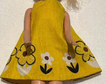 May include: A vintage doll wearing a bright yellow dress with a floral pattern. The dress features large white and yellow flowers with black outlines and green stems. The doll has blonde hair and is posed in a close-up shot.