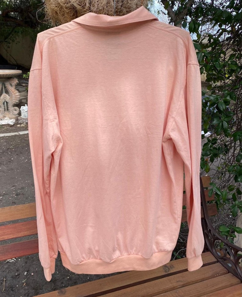 May include: A long-sleeved, light pink collared shirt with a ribbed knit collar and cuffs.
