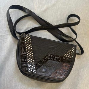 Puede incluir: Un bolso bandolera negro con una correa larga y una solapa curva. El exterior muestra una mezcla de texturas, incluyendo un patrón ondulado, una sección con texto cirílico y un panel con acabado de escamas, acentuado con tachuelas plateadas.