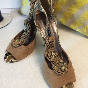 May include: A pair of brown suede heeled sandals with a snakeskin pattern and a flower detail on the front. The sandals have a peep toe and an ankle strap.