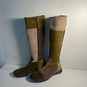 May include: A pair of knee-high boots featuring a patchwork design in shades of olive green, dark brown, and beige. The boots are constructed from suede panels with a vertical zipper closure on the side and a flat, dark-colored rubber sole.