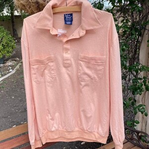 May include: A long-sleeved, peach-colored polo shirt with a button-down collar and two patch pockets. The shirt has a John Blair label on the front.