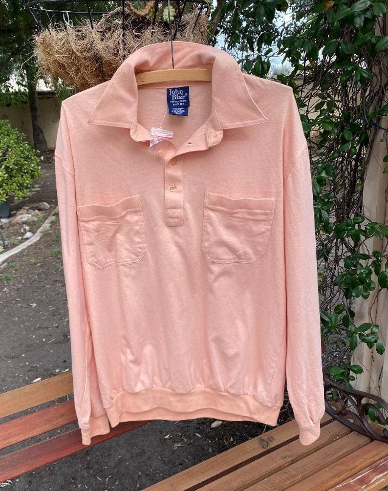 May include: A long-sleeved, peach-colored polo shirt with a button-down collar and two patch pockets. The shirt has a John Blair label on the front.