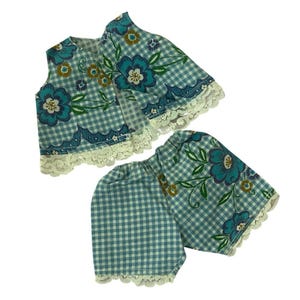 May include: A vintage two-piece outfit featuring a sleeveless top and shorts. The set is made of light blue and white checkered fabric, adorned with large teal floral patterns and white lace trim. The outfit is likely for a child.