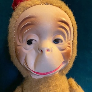 May include: A vintage monkey doll with a painted face, a red felt hat, and a tan plush body. The doll has a smiling expression and is set against a dark teal background. The doll's face has detailed features, including painted eyebrows and a pink mouth.