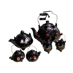 May include: A black ceramic tea set with a rooster design. The set includes a large teapot with a metal handle, four small teapots, and two small teacups. The teapots have red and white accents.