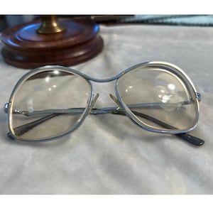Może przedstawiać: Okulary w srebrnej oprawie z przezroczystymi soczewkami. Okulary mają podwójny mostek i klasyczny kształt aviator. Soczewki są lekko przyciemnione. Zausznik jest ciemnoszary.