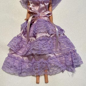 May include: A doll wearing a lavender dress with a satin bodice and a tiered lace skirt. The dress features a matching lace collar and a satin bow at the waist. The doll has blonde hair and is wearing earrings.