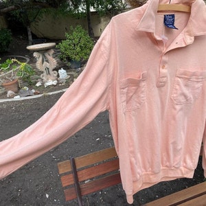 May include: A long-sleeved, peach-colored polo shirt with a button-up collar and two pockets. The shirt has a brand label that reads "Van Heusen" in blue.