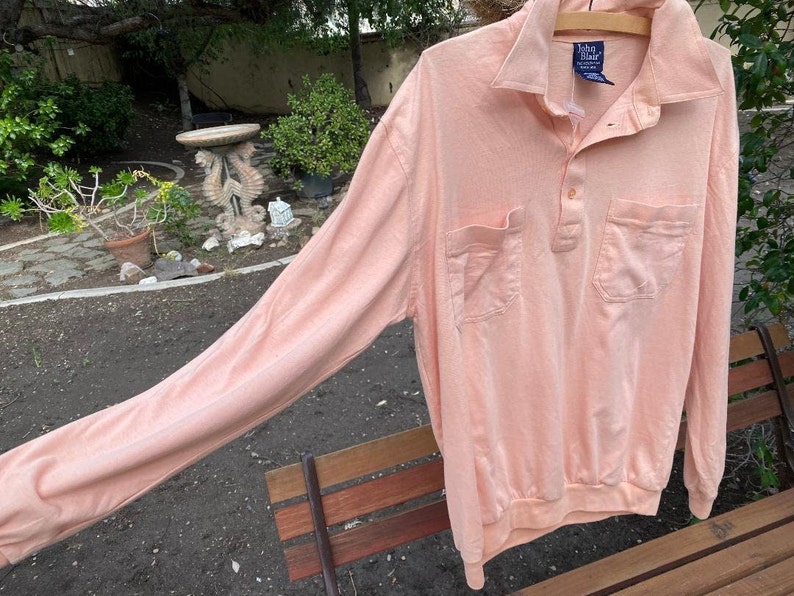 May include: A long-sleeved, peach-colored polo shirt with a button-up collar and two pockets. The shirt has a brand label that reads "Van Heusen" in blue.