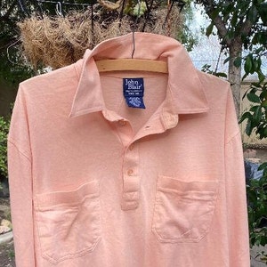 May include: A long-sleeved, light pink collared shirt with a button-up front and two pockets. The shirt has a label that reads "John Blair".