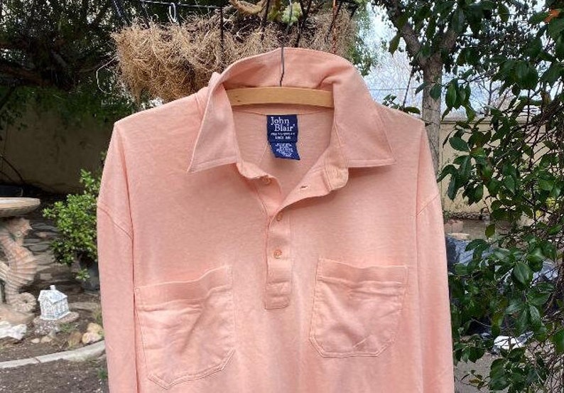May include: A long-sleeved, light pink collared shirt with a button-up front and two pockets. The shirt has a label that reads "John Blair".