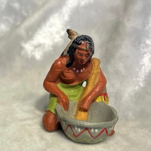 May include: A small hand-painted figurine of a person with dark hair and a feather, wearing a green garment and a white beaded necklace. The figure is kneeling and using a yellow pestle to grind inside a gray bowl decorated with red zig-zag lines.