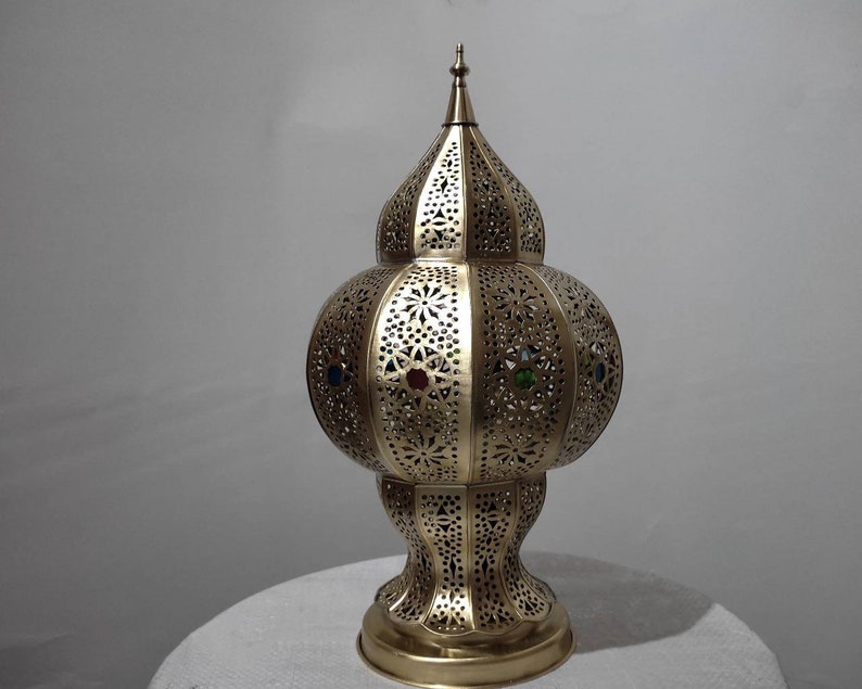 May include: A gold metal lamp with a round, ornate base and a domed top. The lamp has a decorative, cut-out design and features colorful glass accents.