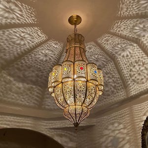 May include: A gold-toned metal pendant light fixture with an intricate laser-cut floral pattern. The lamp features small circular cutouts accented with red, blue, and yellow glass inserts, casting elaborate geometric shadows on the ceiling.