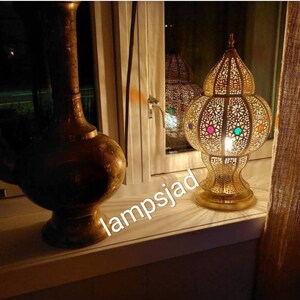 May include: A gold metal table lamp with a round, openwork shade featuring colorful glass accents. The lamp is lit and casting a warm glow. The lamp is sitting on a windowsill with a gold vase in the background.