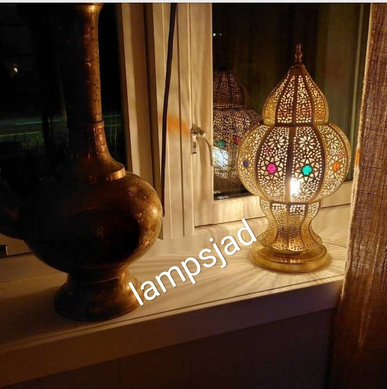 May include: A gold metal table lamp with a round, openwork shade featuring colorful glass accents. The lamp is lit and casting a warm glow. The lamp is sitting on a windowsill with a gold vase in the background.