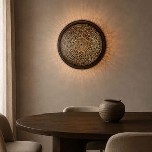May include: A circular wall-mounted light fixture featuring an intricate, laser-cut metal pattern that emits a warm, glowing light. The design creates a radial, sunburst-like shadow effect on the surrounding wall surface, highlighting the detailed geometric cutouts.