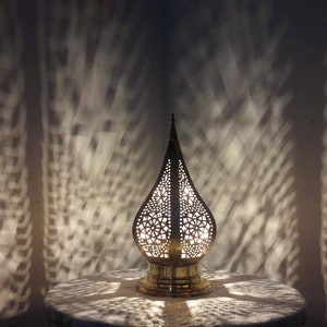May include: A gold metal table lamp with a decorative, intricate, cut-out design in the shape of a teardrop. The lamp is lit and casting a pattern of light on the white surface below.