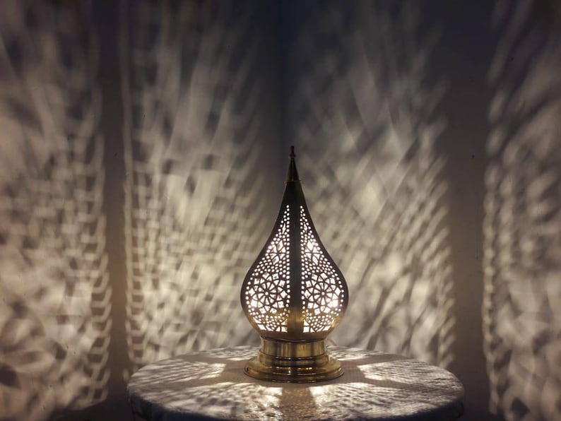 May include: A gold metal table lamp with a decorative, intricate, cut-out design in the shape of a teardrop. The lamp is lit and casting a pattern of light on the white surface below.