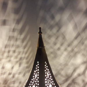May include: A gold metal lamp with a pointed top and intricate cut-out designs. The lamp casts a shadow on the wall behind it.