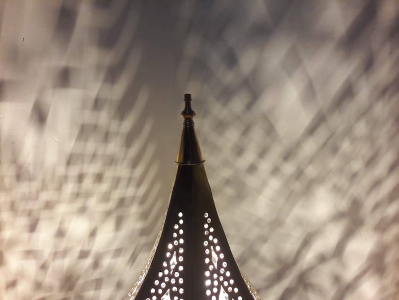 May include: A gold metal lamp with a pointed top and intricate cut-out designs. The lamp casts a shadow on the wall behind it.