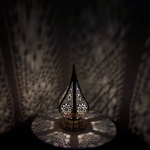 May include: A gold metal lantern with intricate cut-out designs casts a patterned shadow on a white surface. The lantern has a teardrop shape and a small base.