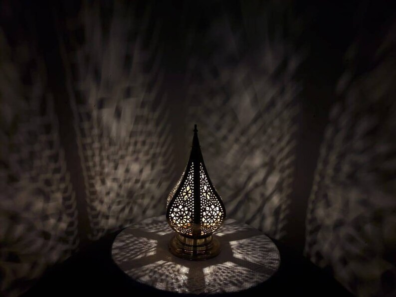 May include: A gold metal lantern with intricate cut-out designs casts a patterned shadow on a white surface. The lantern has a teardrop shape and a small base.