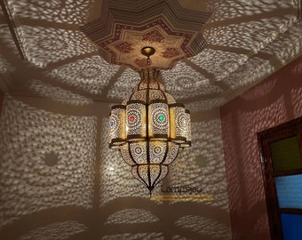 May include: A large, ornate brass chandelier with intricate cutouts and colored glass accents hangs from a ceiling with a patterned design. The chandelier casts intricate shadows on the wall.