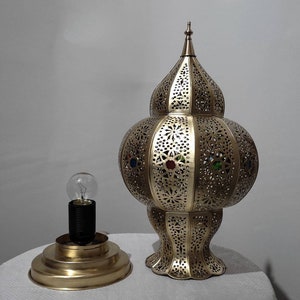 May include: A gold metal lamp with a decorative, intricate design and colorful glass accents. The lamp has a round base and a tall, pointed top.