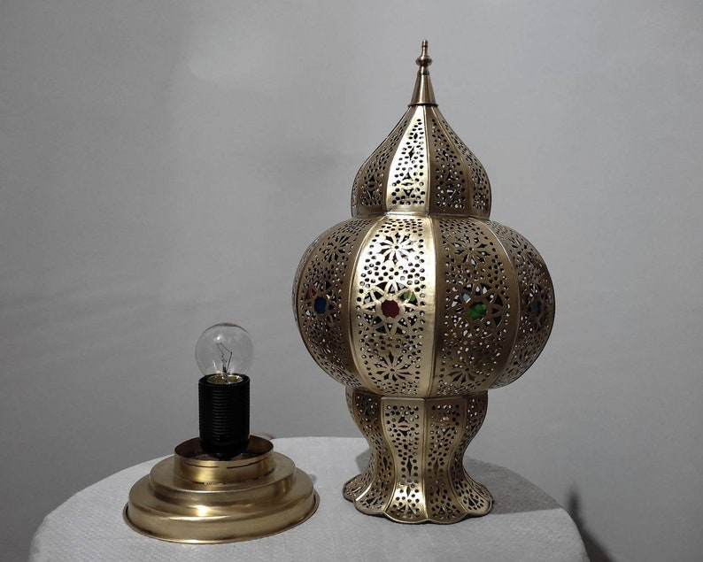 May include: A gold metal lamp with a decorative, intricate design and colorful glass accents. The lamp has a round base and a tall, pointed top.