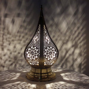 May include: A brass table lamp with a decorative, intricate, cut-out design in the shape of a teardrop. The lamp is lit and casting a shadow on the table.