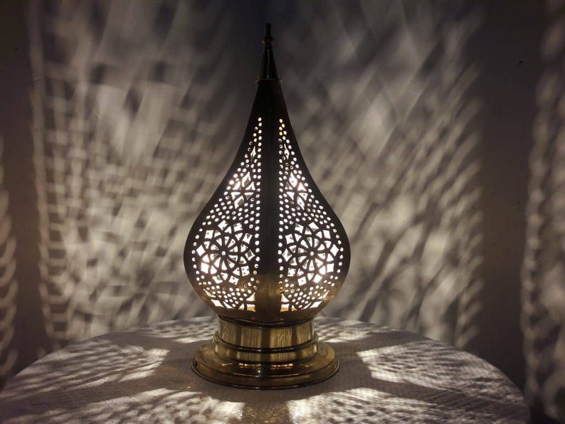 May include: A brass table lamp with a decorative, intricate, cut-out design in the shape of a teardrop. The lamp is lit and casting a shadow on the table.