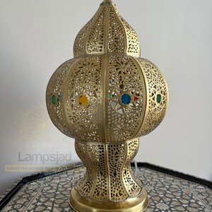 May include: A gold Moroccan lantern with intricate cutouts and colorful glass accents. The lantern has a round body and a pointed top.