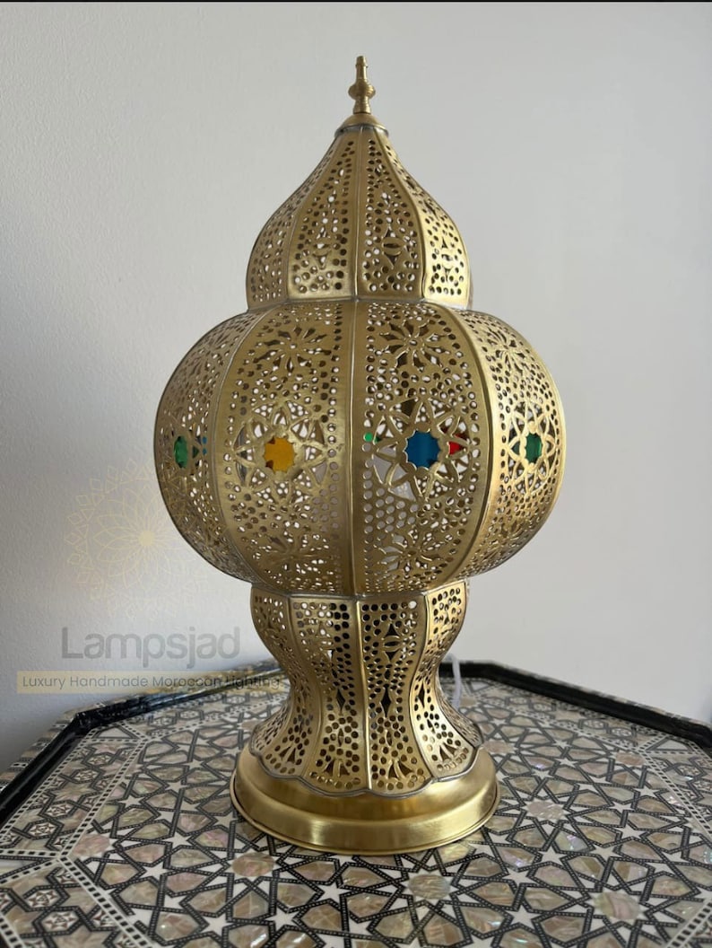 May include: A gold Moroccan lantern with intricate cutouts and colorful glass accents. The lantern has a round body and a pointed top.
