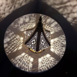 May include: A white metal lantern with intricate cut-out designs casts a patterned shadow on a white surface. The lantern has a pointed top and a gold base.