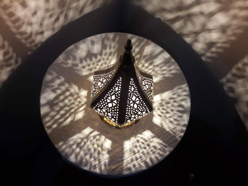 May include: A white metal lantern with intricate cut-out designs casts a patterned shadow on a white surface. The lantern has a pointed top and a gold base.