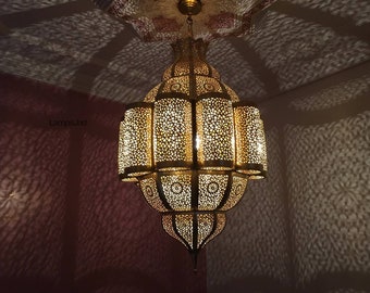 May include: A large, ornate, gold-colored metal chandelier with intricate cut-out designs. The chandelier is hanging from a ceiling and is illuminated, casting shadows on the wall.