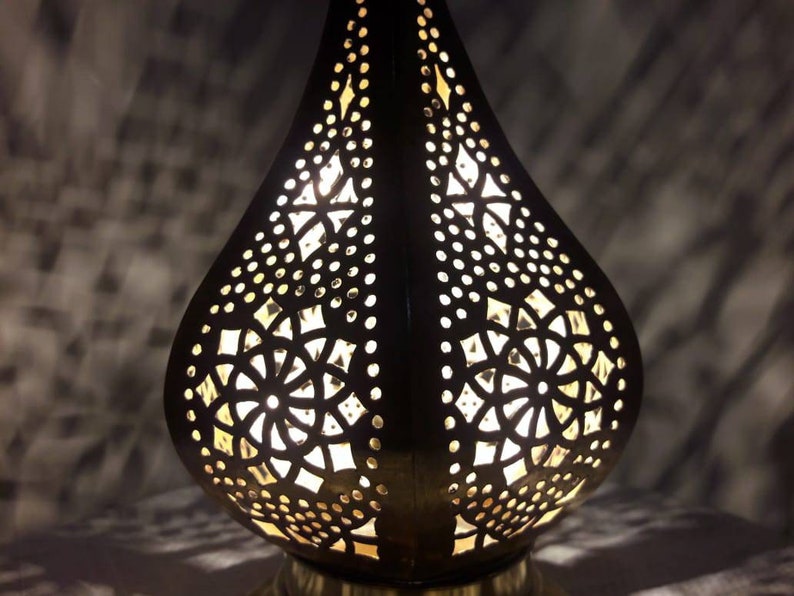 May include: A black metal lamp with intricate cut-out designs in the shape of a teardrop. The lamp is illuminated from within, casting a warm glow through the intricate patterns.
